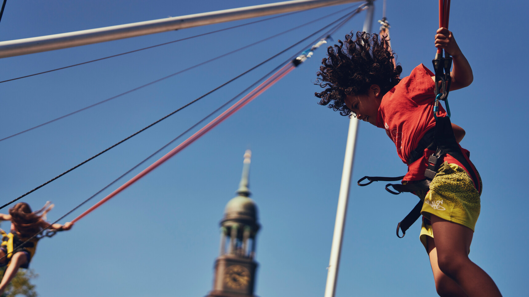 Kind springt mit Gurt an einem Trampolin bei klarem Himmel vor einem Kirchturm.