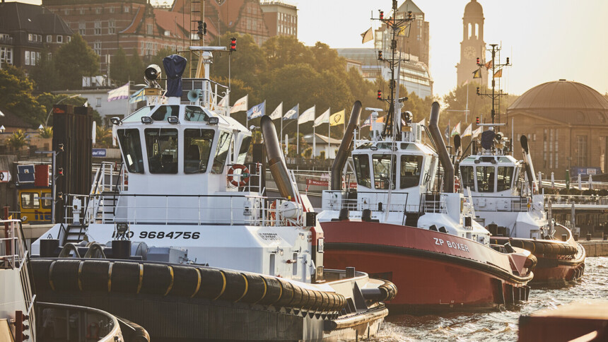 Drei Schlepper liegen im Hafen vor einer Stadt mit historischen Gebäuden und Fahnen.