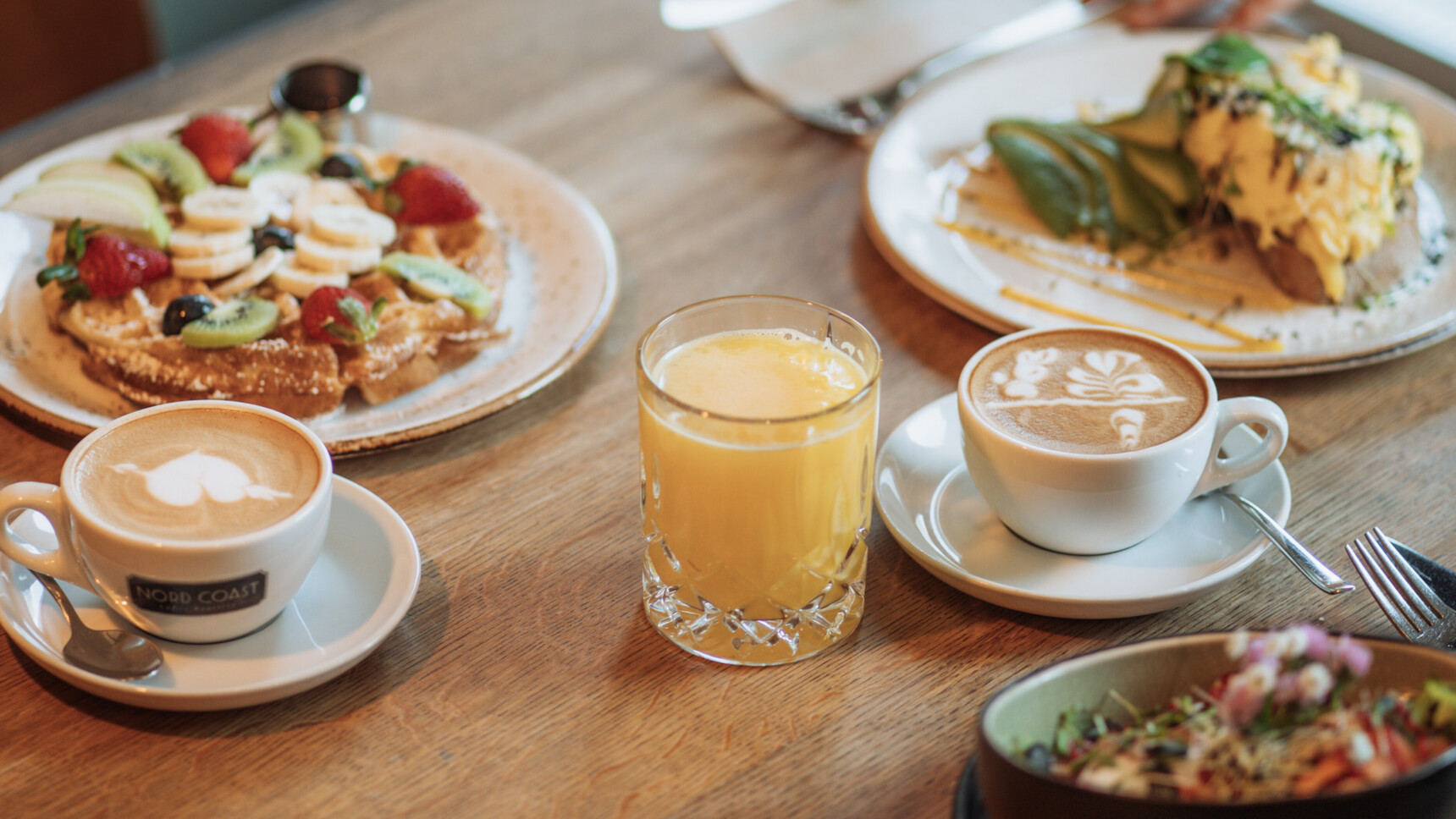Frühstückstisch mit Kaffee, Orangensaft, Waffeln mit Früchten und herzhaftem Gericht auf Holztisch.