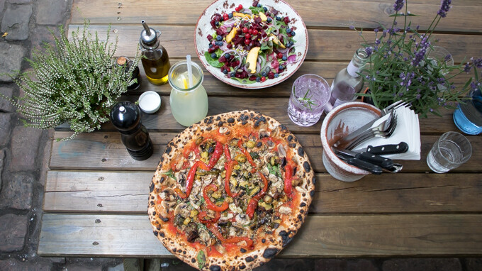 Pizza mit Gemüse, Salat, Getränke und Gewürze auf einem Holztisch im Freien.