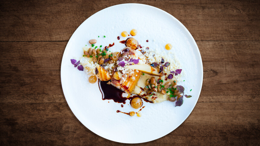 Gourmetgericht mit Fleisch, Sauce, Bohnen, Kräutern und Püree auf weißem Teller vor Holzhintergrund.