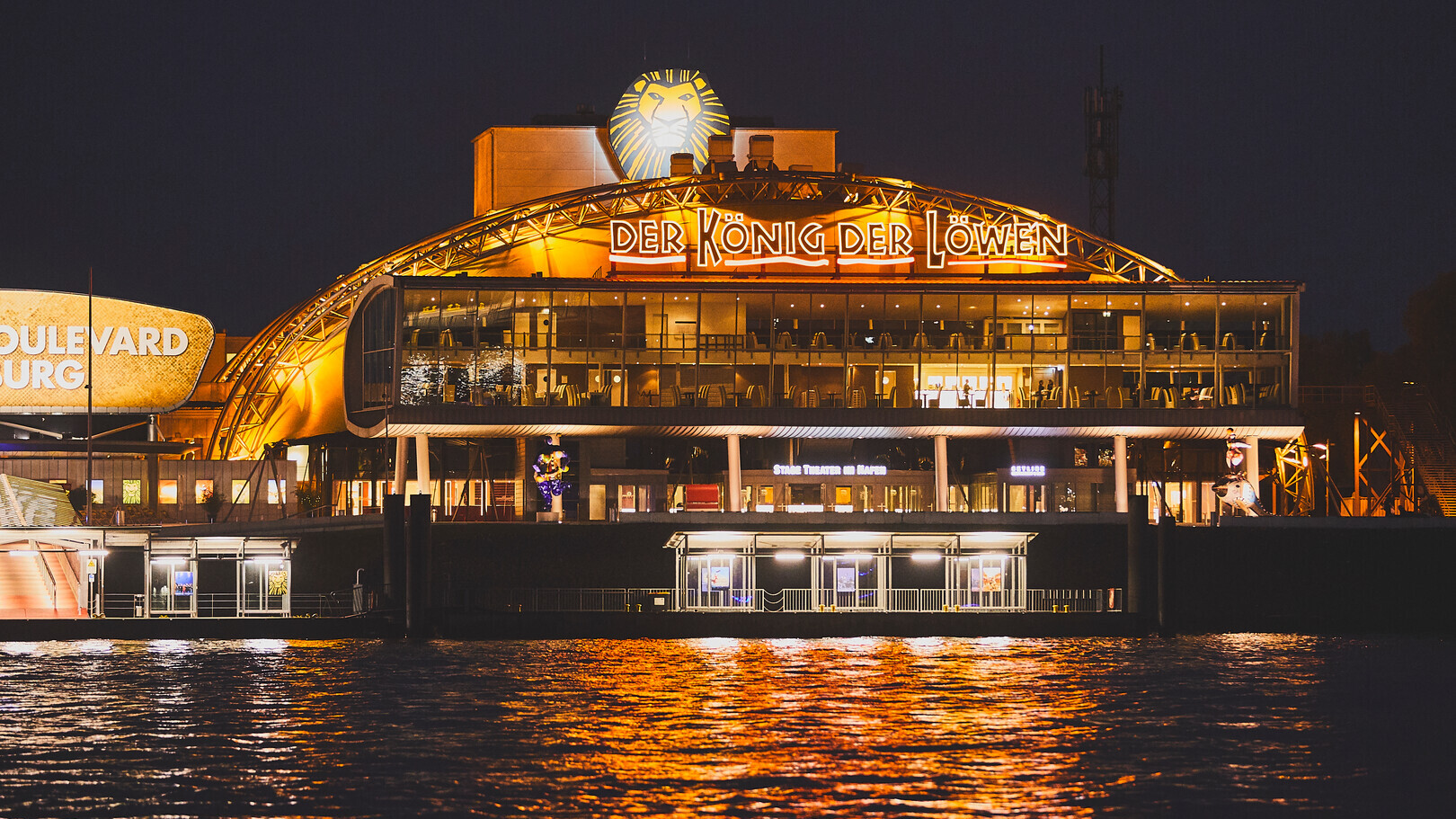 Beleuchtetes Theatergebäude „Der König der Löwen“ am Wasser bei Nacht in Hamburg.