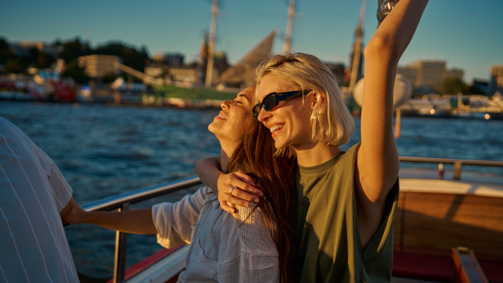 Zwei lachende Frauen umarmen sich glücklich auf einem Boot bei Sonnenuntergang am Wasser.