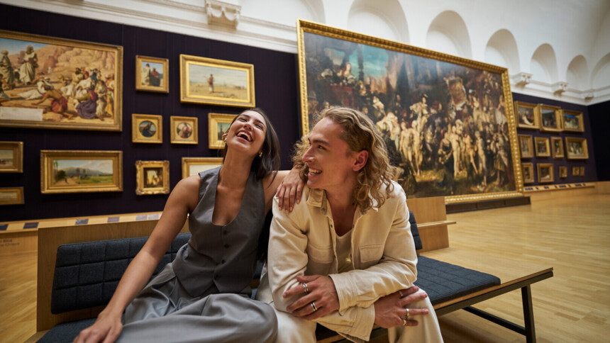 Zwei junge Menschen sitzen lachend auf einer Bank in der Hamburger Kunsthalle. Sie wirken entspannt und glücklich, während sie von beeindruckenden Gemälden in einem großen, hellen Ausstellungsraum umgeben sind.