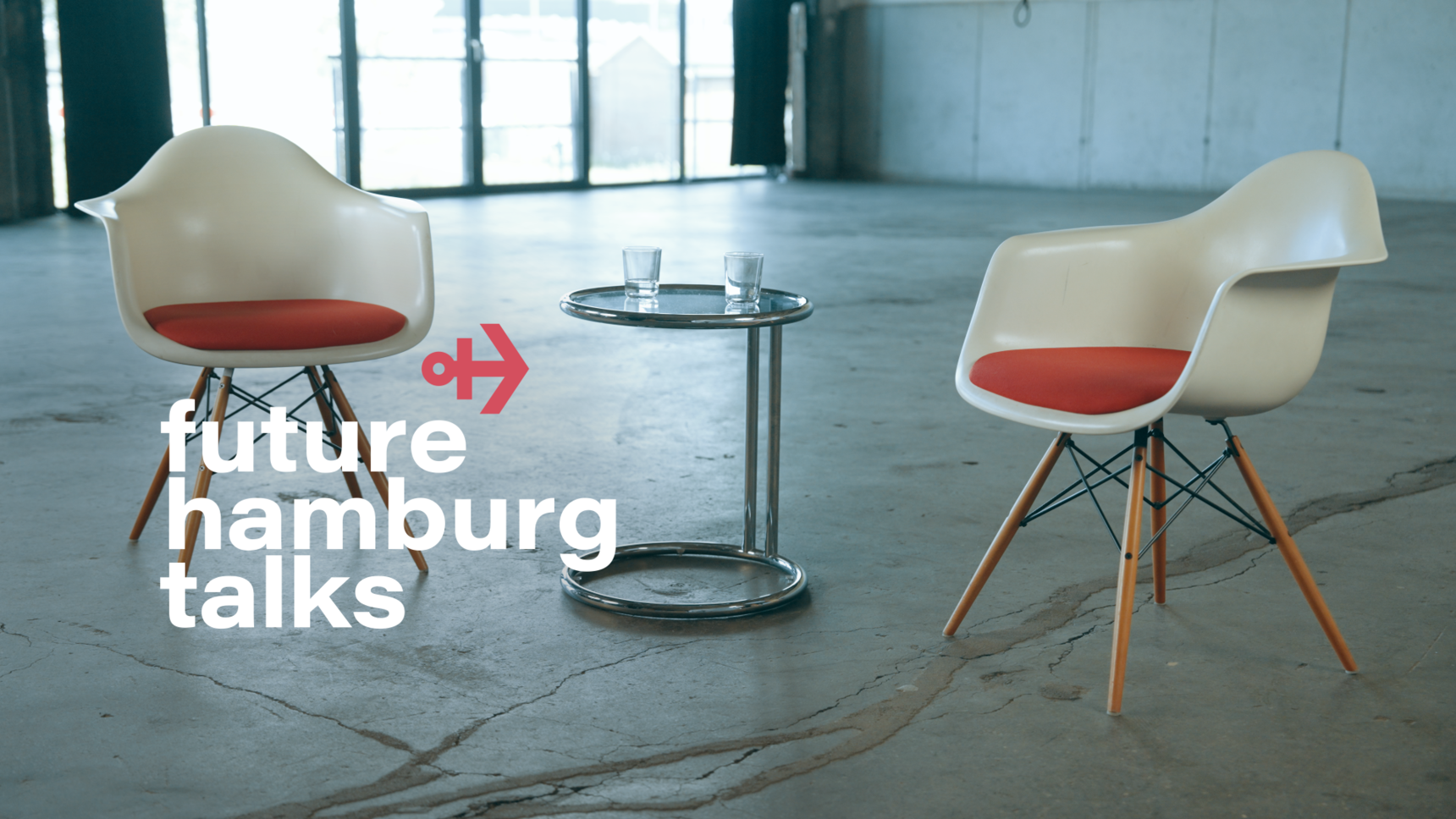 Zwei weiße Stühle mit roten Sitzpolstern stehen sich in einem offenen Raum gegenüber. Dazwischen ein runder Glastisch mit zwei Wassergläsern. Links im Bild das Future Hamburg Talks-Logo mit einem roten Pfeilsymbol.