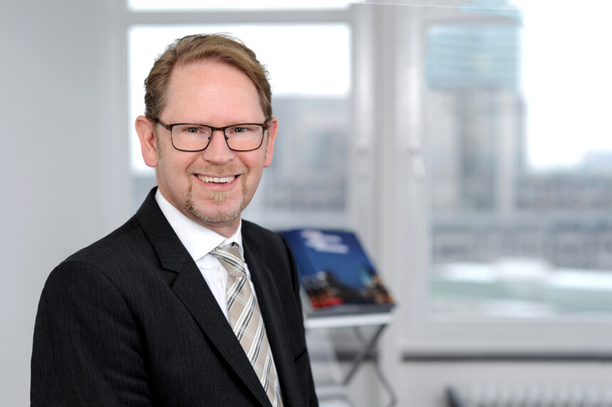 Porträt eines lächelnden Mannes in Anzug und Brille vor einem Fenster mit Stadtblick.