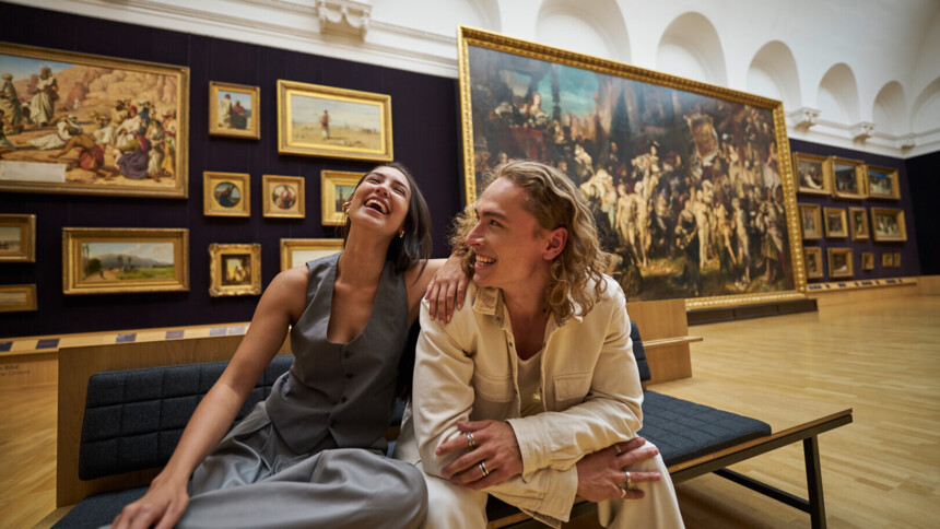 Zwei junge Menschen sitzen lachend auf einer Bank in der Hamburger Kunsthalle. Sie wirken entspannt und glücklich, während sie von beeindruckenden Gemälden in einem großen, hellen Ausstellungsraum umgeben sind.