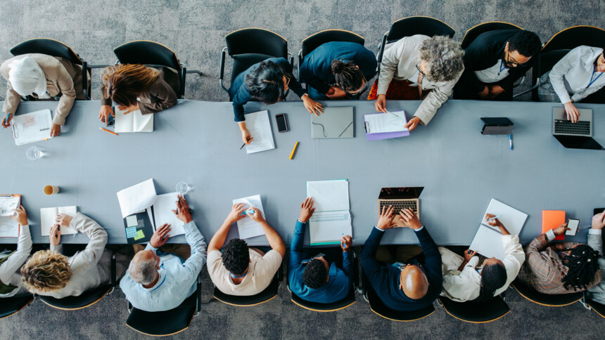Verschiedene Menschen sitzen an einem langen Tisch und arbeiten gemeinsam an Dokumenten und Laptops.
