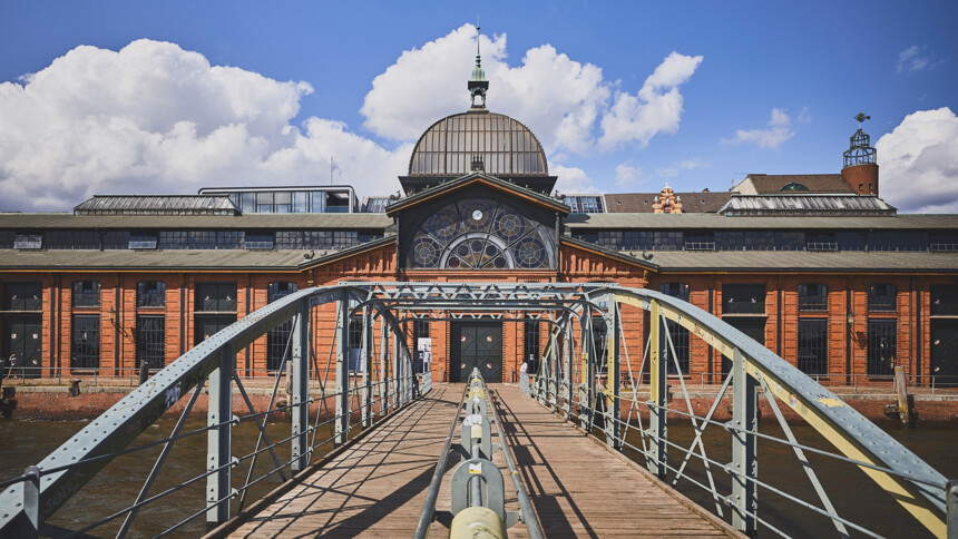 Blick über eine Stahlbrücke auf ein historisches Backsteingebäude mit großen Fenstern und einer Kuppel.