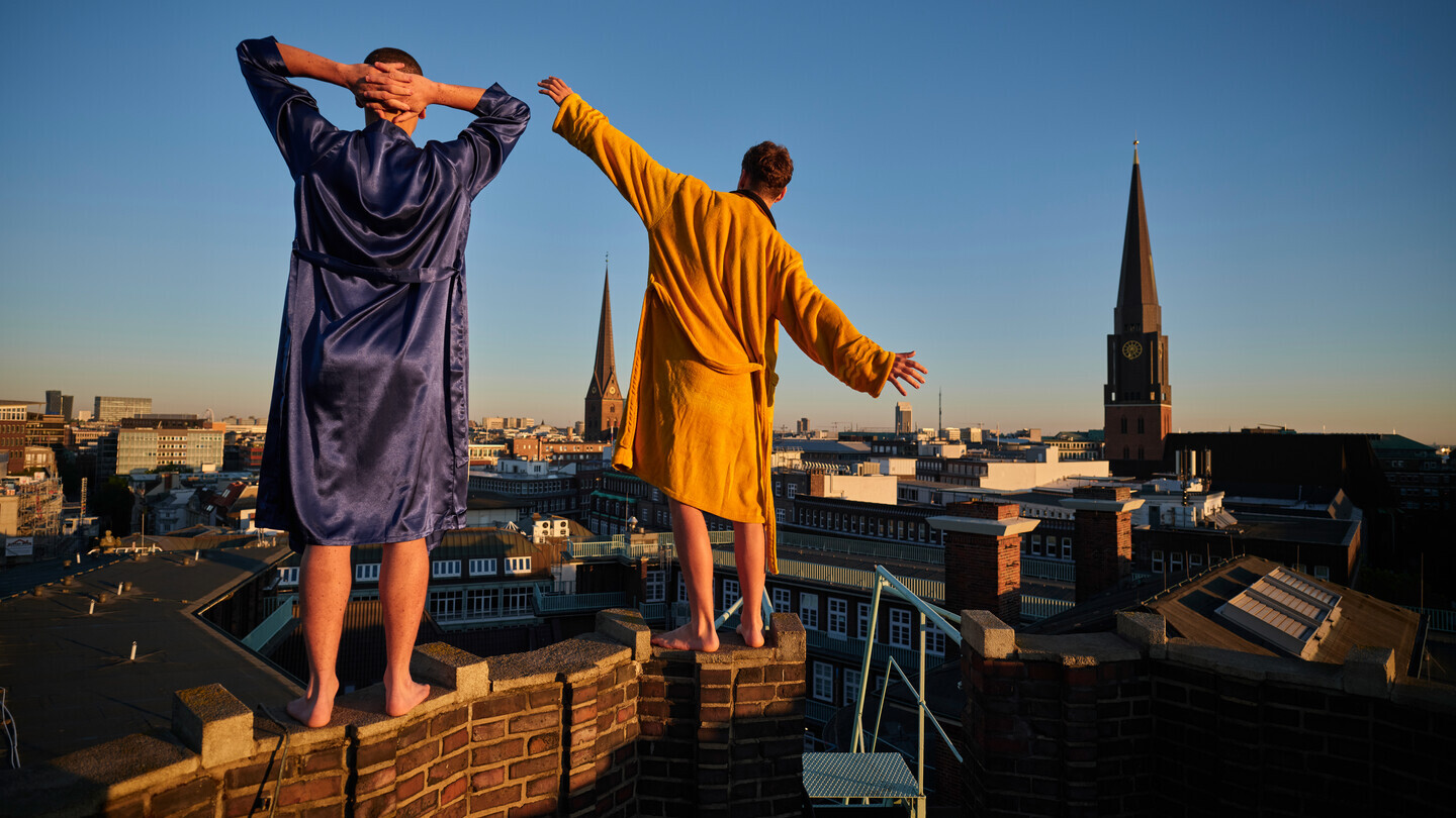 Zwei Männer in Bademänteln stehen auf einer Mauer und blicken über eine Stadt bei Sonnenuntergang.