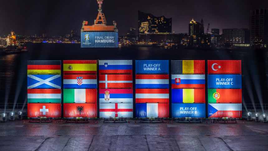 Nachtaufnahme des Hamburger Hafens mit beleuchteten Schiffscontainern, die die Flaggen der Teilnehmerländer der UEFA EURO 2024 zeigen. Ein Kran hebt einen Container mit der Aufschrift 'Final Draw Hamburg'. Im Hintergrund ist die Elbphilharmonie und die Skyline von Hamburg zu sehen.