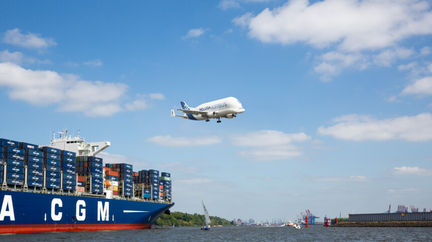Ein Airbus Beluga fliegt über einen Containerfrachter im Hafen bei blauem Himmel.