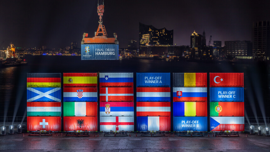 Nachtaufnahme des Hamburger Hafens mit beleuchteten Schiffscontainern, die die Flaggen der Teilnehmerländer der UEFA EURO 2024 zeigen. Ein Kran hebt einen Container mit der Aufschrift 'Final Draw Hamburg'. Im Hintergrund ist die Elbphilharmonie und die Skyline von Hamburg zu sehen.