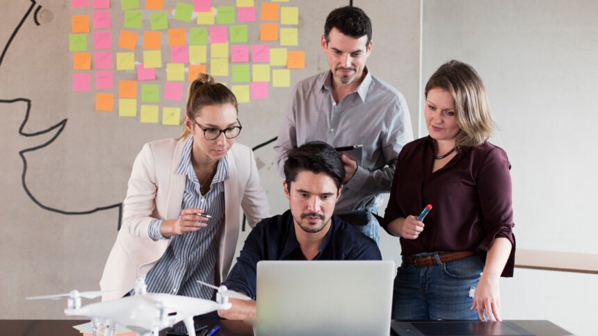 Vier Personen arbeiten gemeinsam an einem Laptop, mit Drohne und bunten Haftnotizen im Hintergrund.