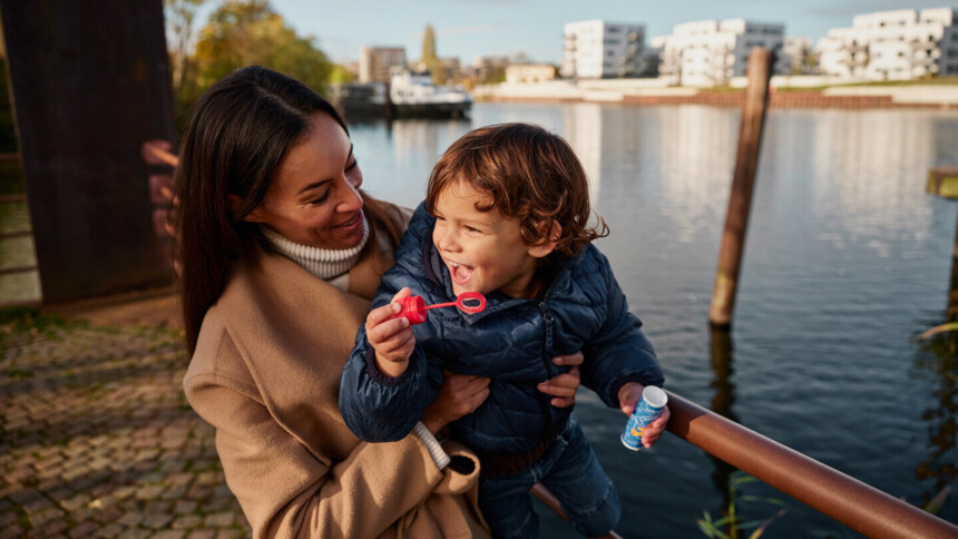 Frau hält lachendes Kind am Flussufer, das Seifenblasen macht.